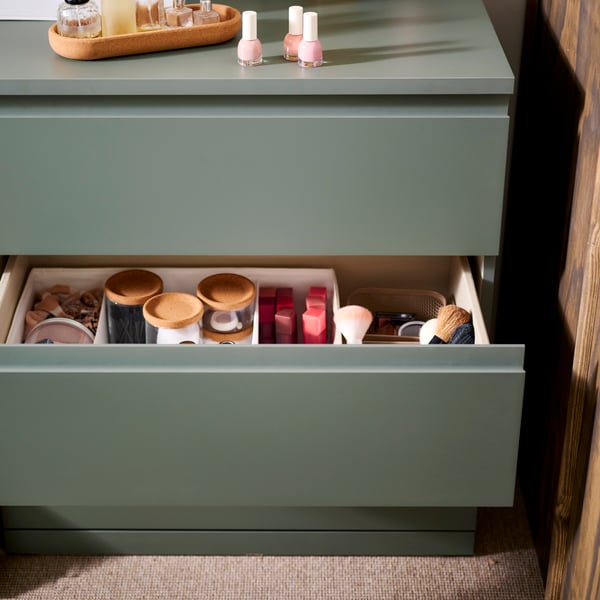 Une commode à 3 tiroirs STORKLINTA gris-vert positionnée à côté d’un mur brun, son tiroir central ouvert laissant entrevoir des boîtes et autres contenants.