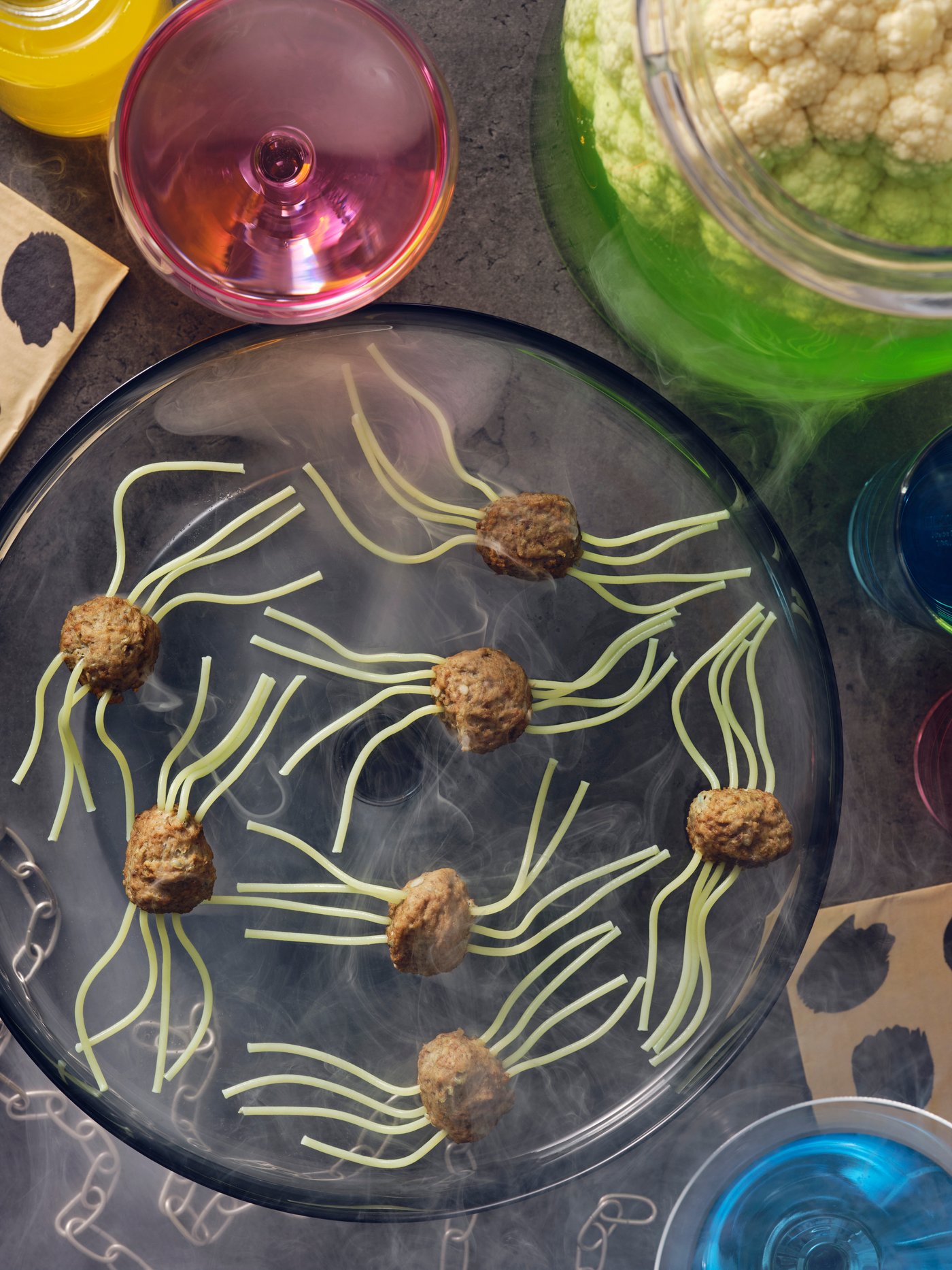 A plate of HUVUDROLL plant balls has green pasta tendrils making them look like spiders, surrounded by colourful drinks.