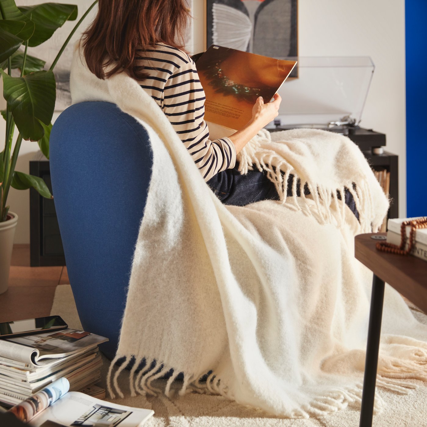 Une personne assise sur un fauteuil relax pivotant LILLESÄTER au tissu Gunnared bleu foncé avec un jeté KLIPPOXEL doux, à côté de quelques magazines.