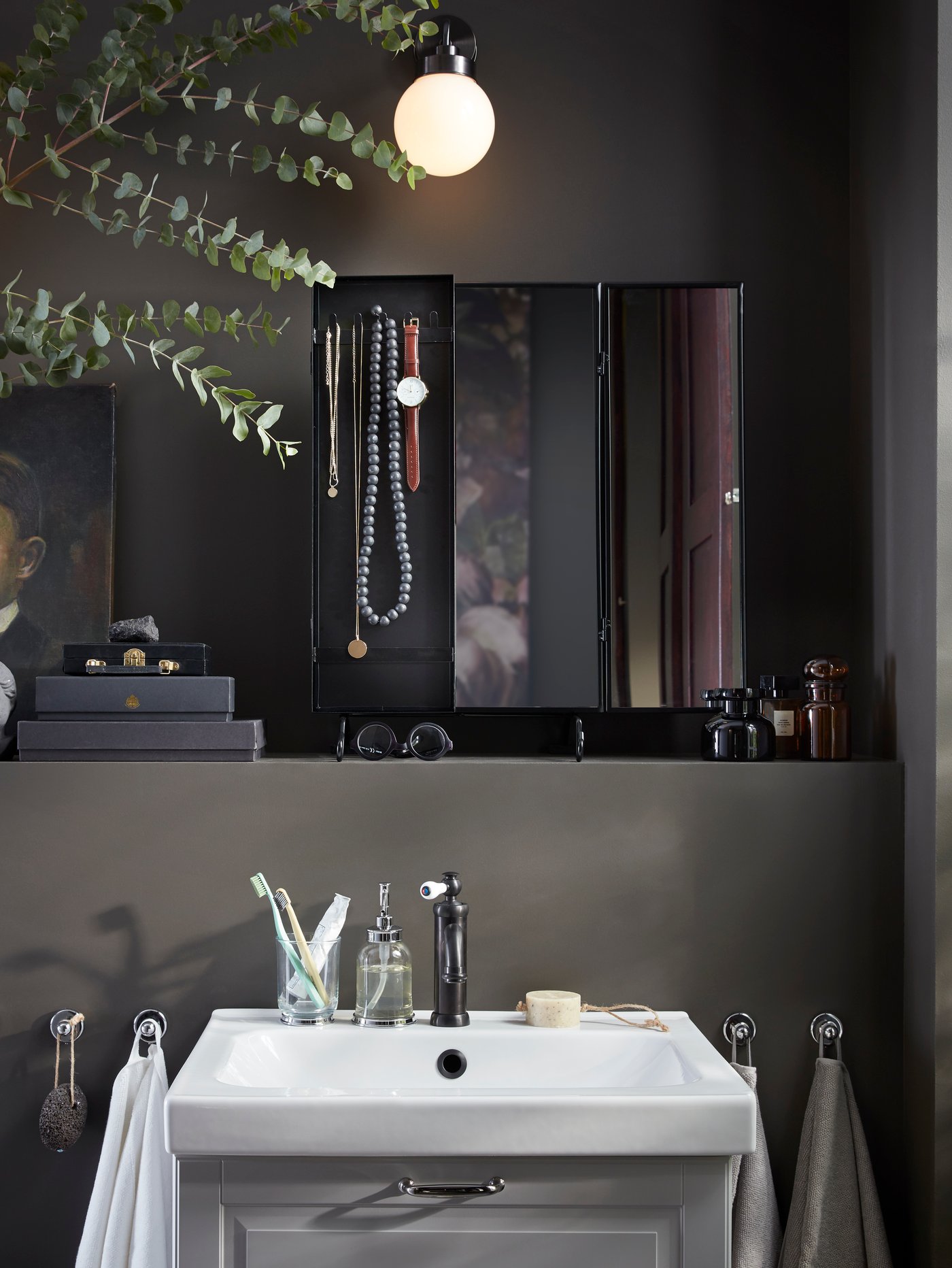 A dark grey bathroom with a black mirror on a shelf above a wash-basin, jewellery and accessories hang from the mirror.