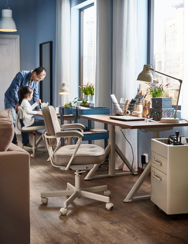 A man and a child in a room with IDÅSEN desks in ash veneer next to the windows, HEKTAR work lamps and beige office chairs