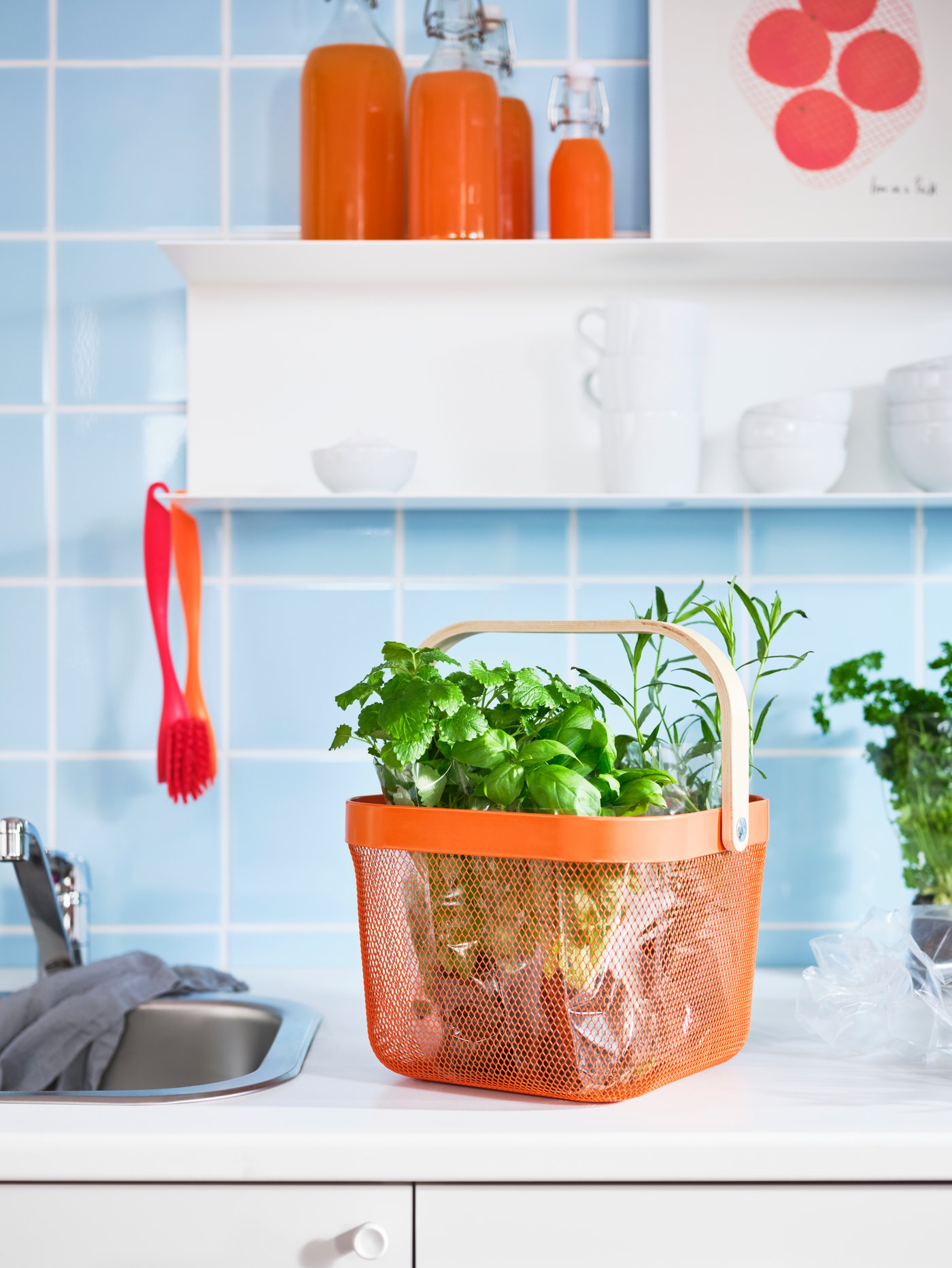 A kitchen top with herbs in an orange RISATORP basket. A white shelf on the kitchen wall holds cups, bowls and juice bottles.