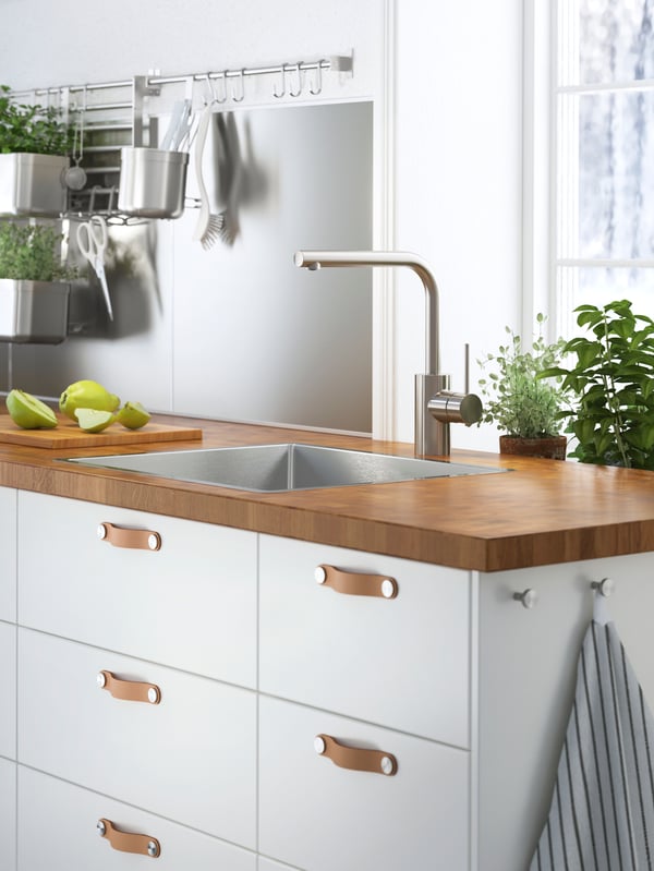 A worktop in thick oak veneer on a cabinet with white fronts and leather handles. A sink and a cutting board with pears.