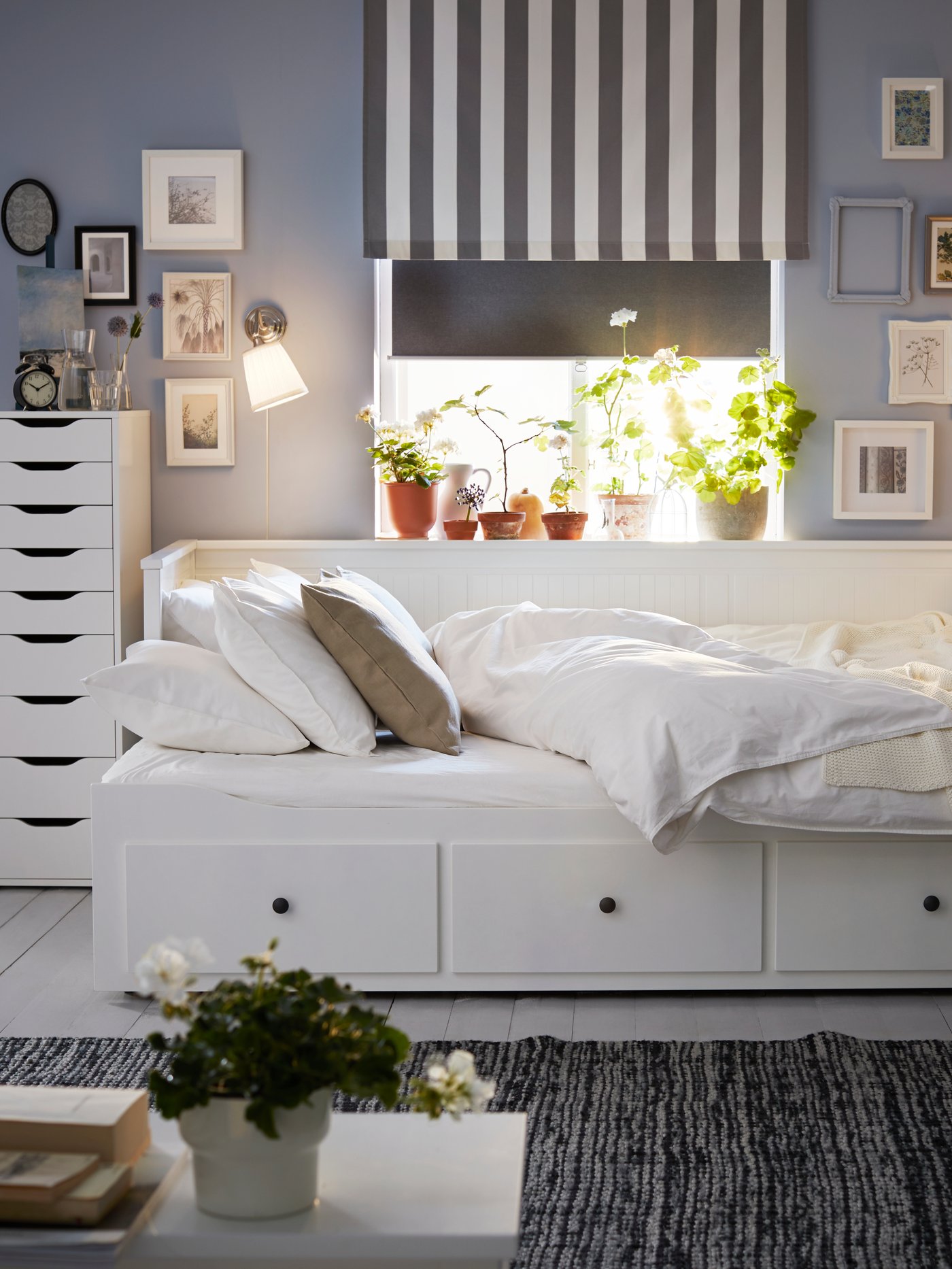 A HEMNES daybed with white quilt cover and pillowcases in front of a window with a grey roller blind half-way down.