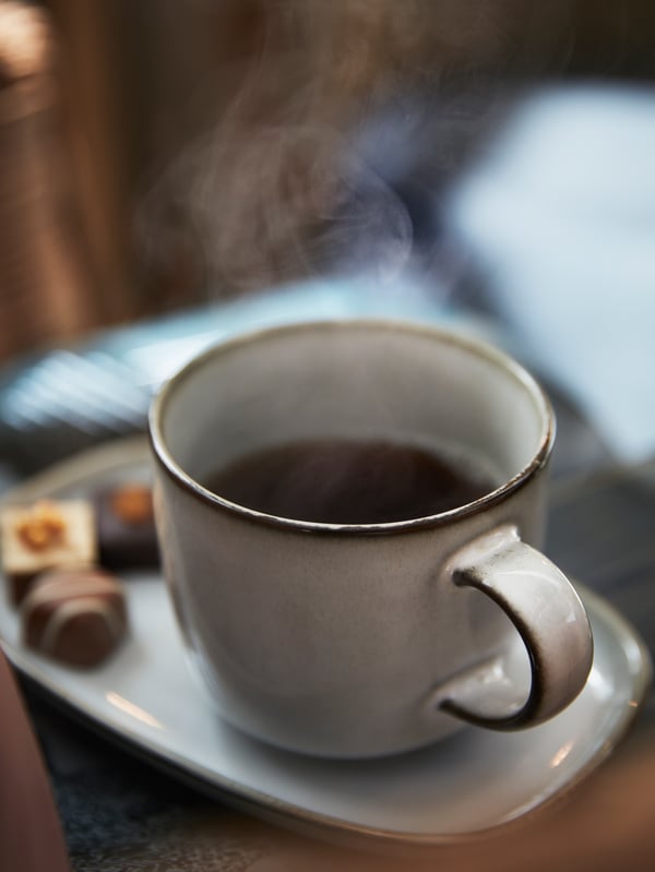 A grey GLADELIG mug holding hot steaming coffee placed on a grey plate with three chocolate pieces.