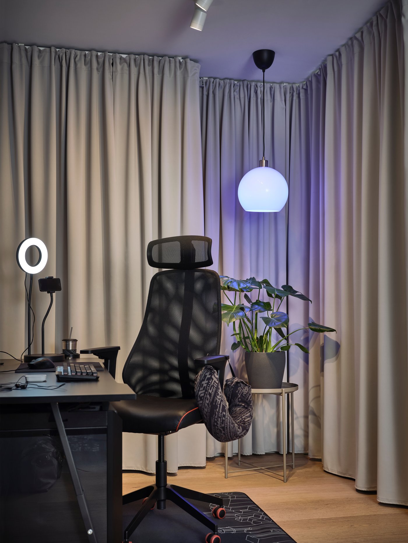 A black MATCHSPEL chair and UTESPELARE desk with a ring light on top in a curtained area with a lamp and a plant behind it.