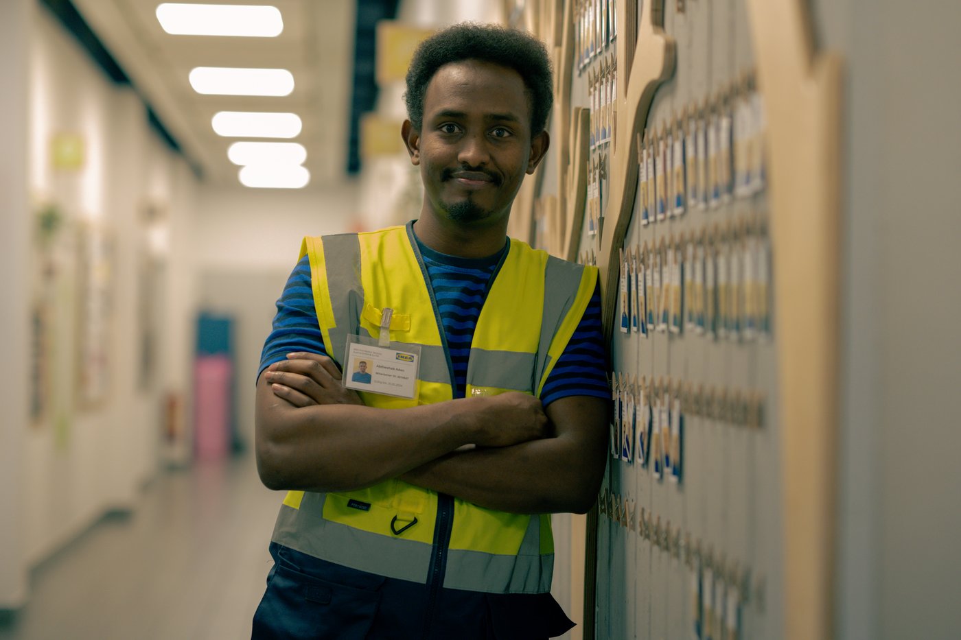 Abdi, wearing a high-visibility vest, stands with arms crossed in a corridor, smiling.