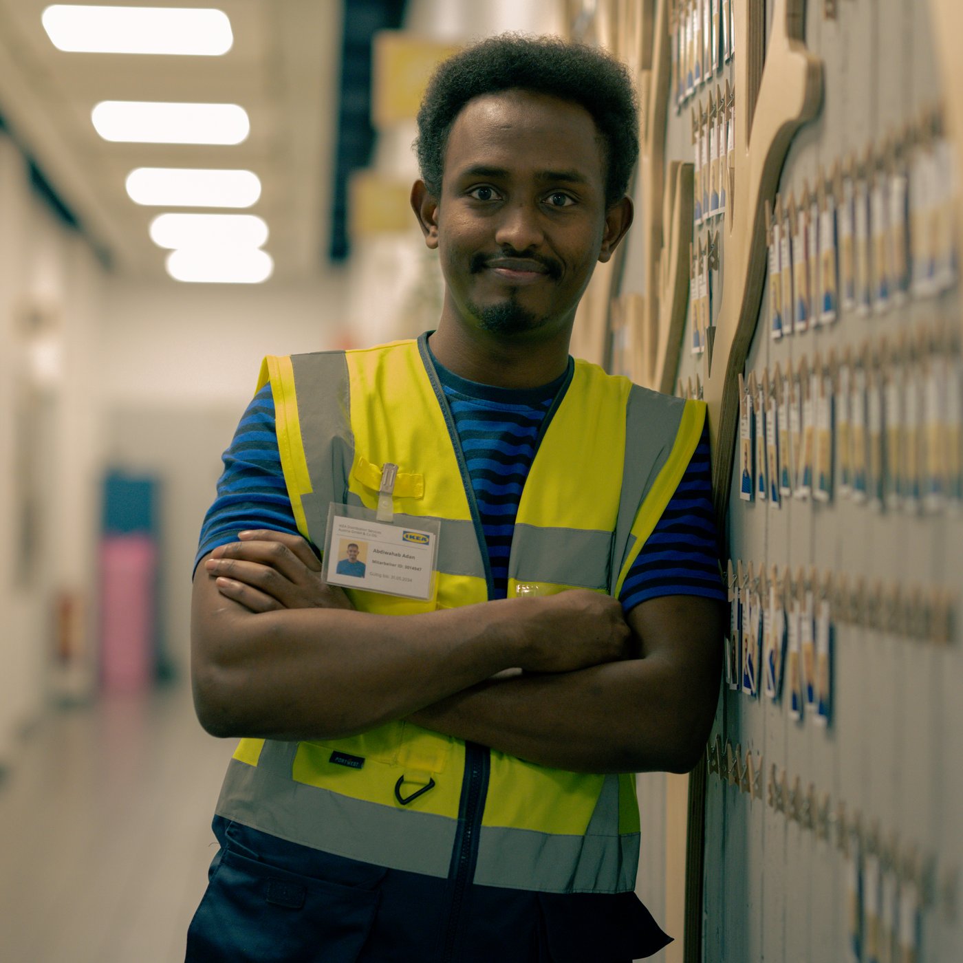 Abdi, der eine Warnweste trägt, steht mit verschränkten Armen in einem Korridor und lächelt.
