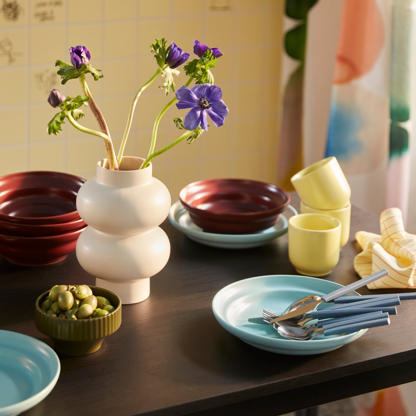 A dining table set with a SPJUTLILJA 3-piece serving set, including a dark purple bowl, a yellow cup and a light blue plate.