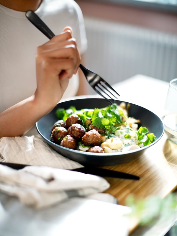 A person in a white t-shirt holds a fork over a dish of HUVUDROLL plant balls, potatoes, sauce and fresh herbs.