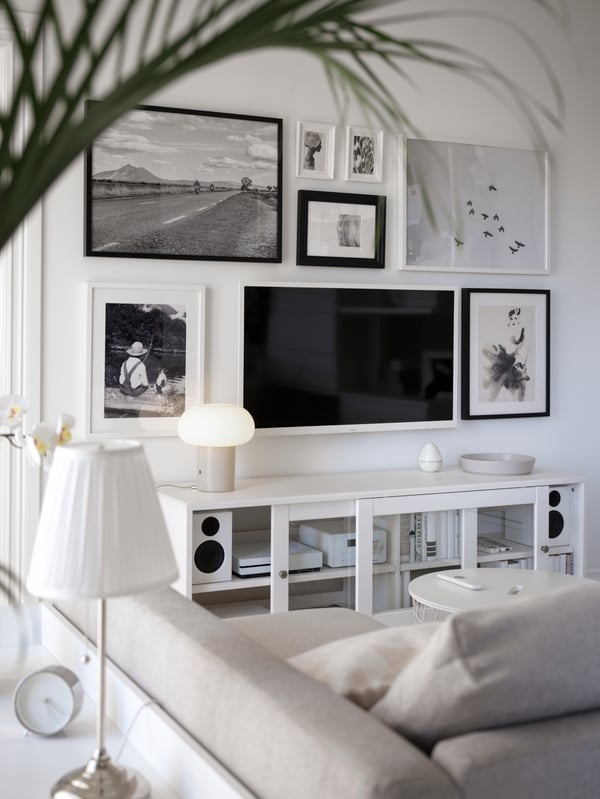 A wall-mounted TV hangs amid framed art over a white HAVSTA TV bench opposite a beige FRIHETEN sofa-bed in a living room.