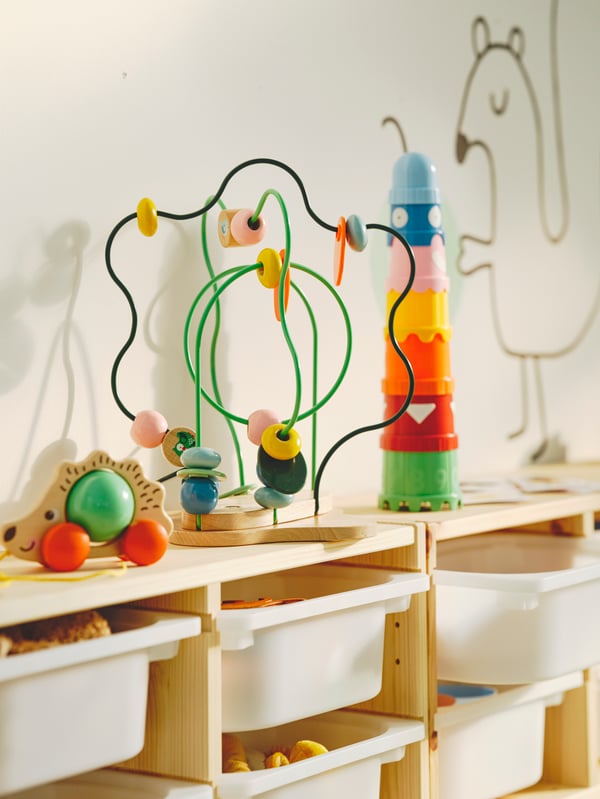 An UPPSTÅ bead maze toy between stacking cups and a hedgehog toy on top of a TROFAST frame with white storage boxes in it.