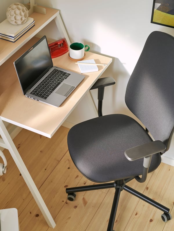 A small corner workspace with a MITTPLAN desk, a dark grey SMÖRKULL office chair, a laptop, and a white and green coffee cup.