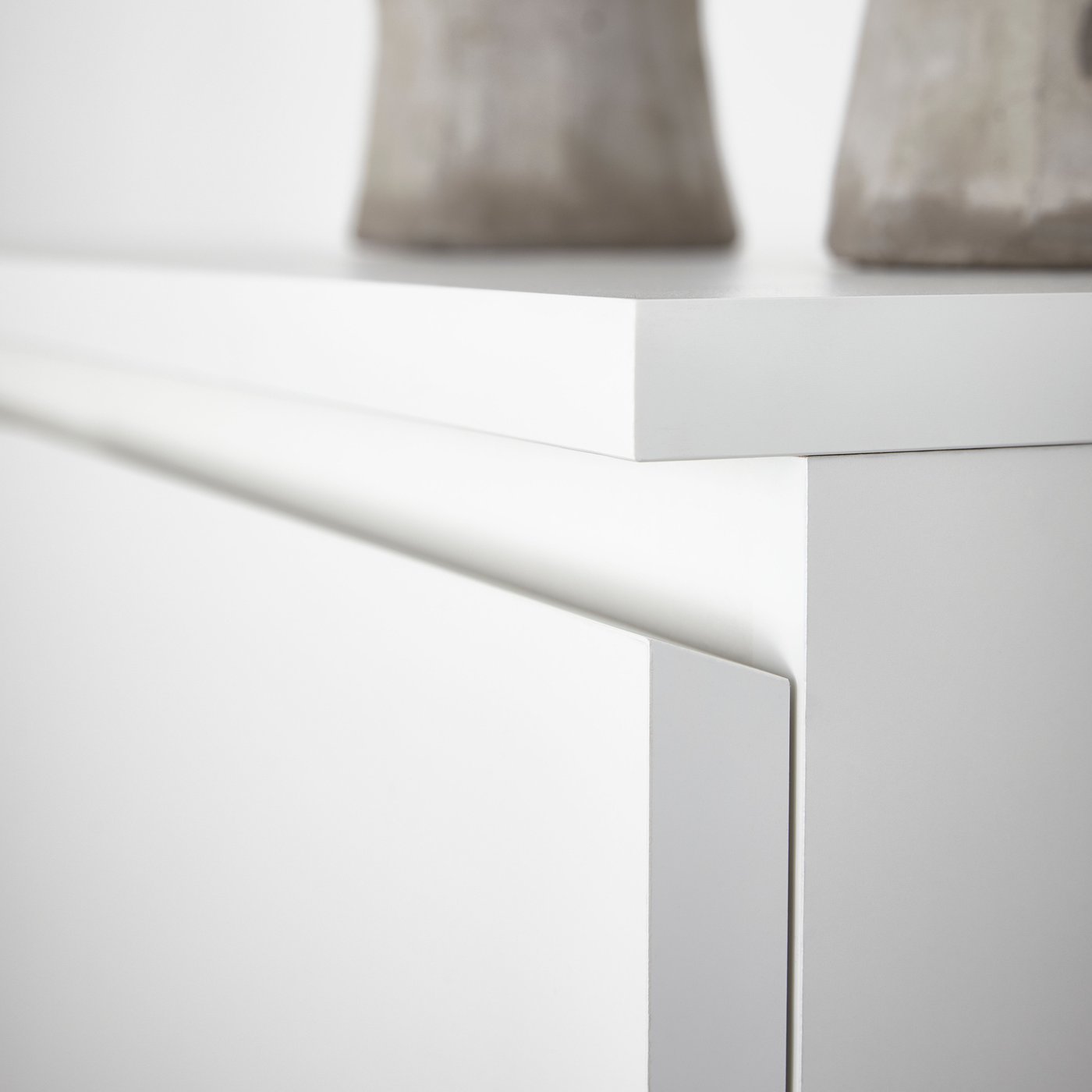 The corner of the surface and the top drawer of a white MALM chest with 2 drawers with two light grey objects on top of it.