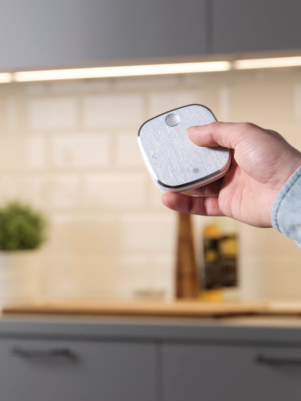 En primer plano se ve una mano con un control para iluminación inteligente STYRBAR apuntando hacia luces LED integradas en un mueble de cocina.