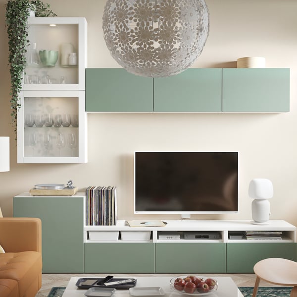 The wall in a living room has white BESTÅ wall and media storage with gray-green fronts by a TV and a lamp.