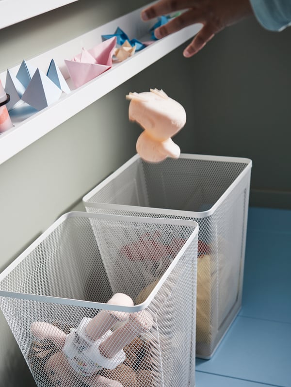 A picture ledge, two white DRÖNJÖNS wastepaper baskets with toys inside and a boy’s hand throwing a toy into one of them.