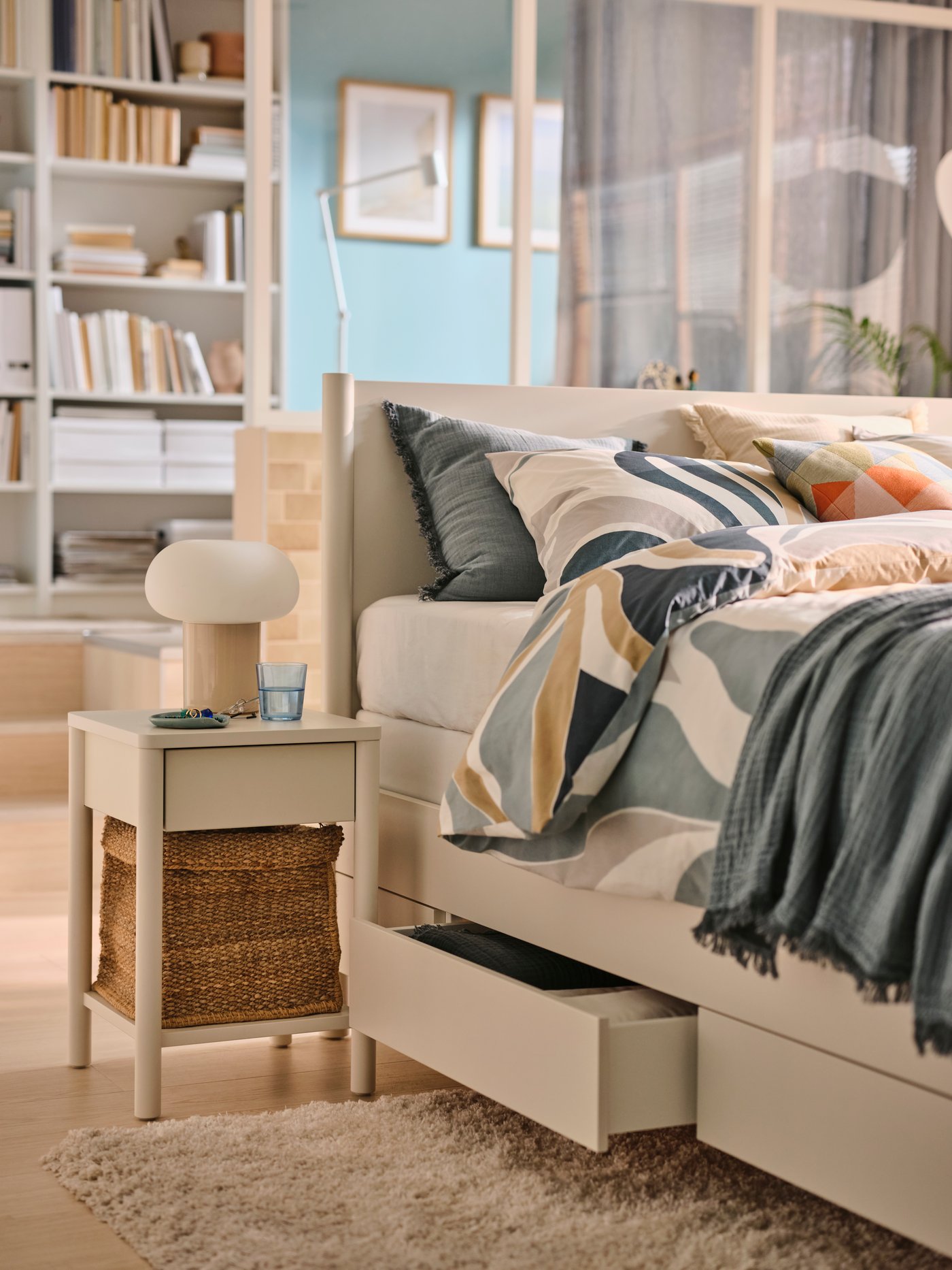 An off-white TONSTAD bedside table beside an off-white TONSTAD bed covered in MYGGLASVINGE multicolour bed linen.