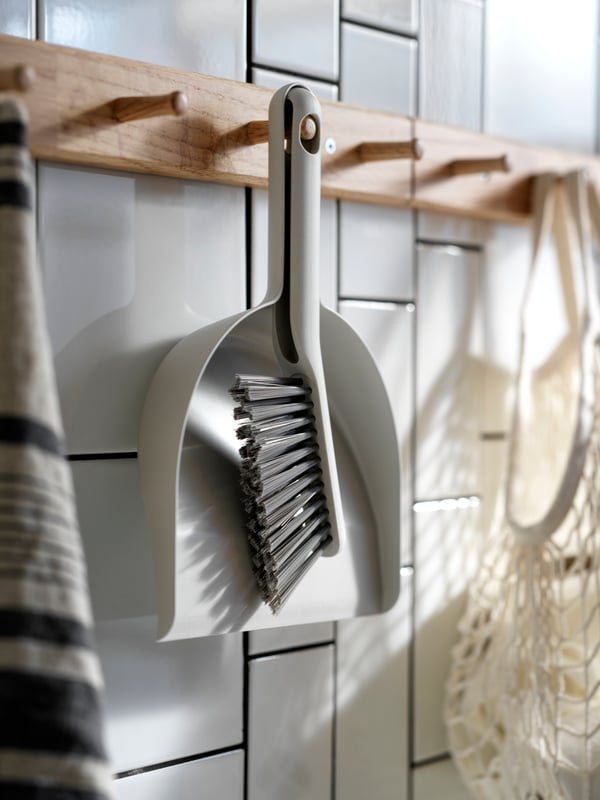 A PEPPRIG dustpan and brush in grey hanging from a wooden oak rack along with other objects on a tiled wall.