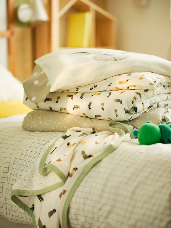 Gros plan sur des textiles de lit DRÖMSLOTT pliés sur un lit d’enfant avec housse de couette, à côté d’une chenille en peluche.
