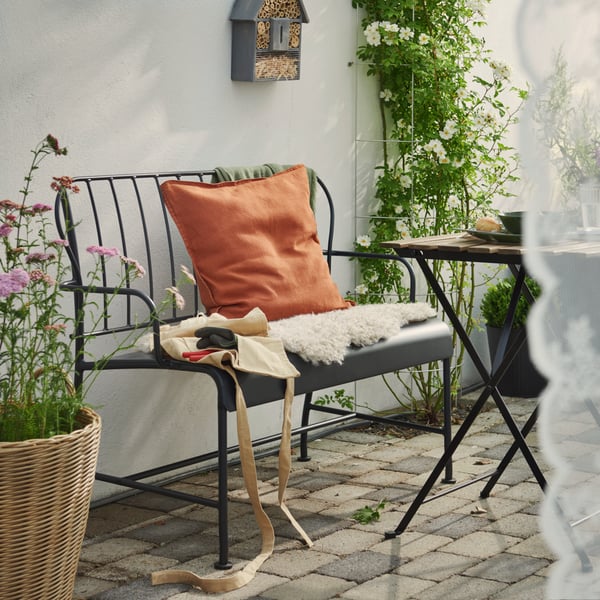 A patio featuring a grey LÄCKÖ outdoor sofa with cushions, surrounded by green plants and a stone floor.