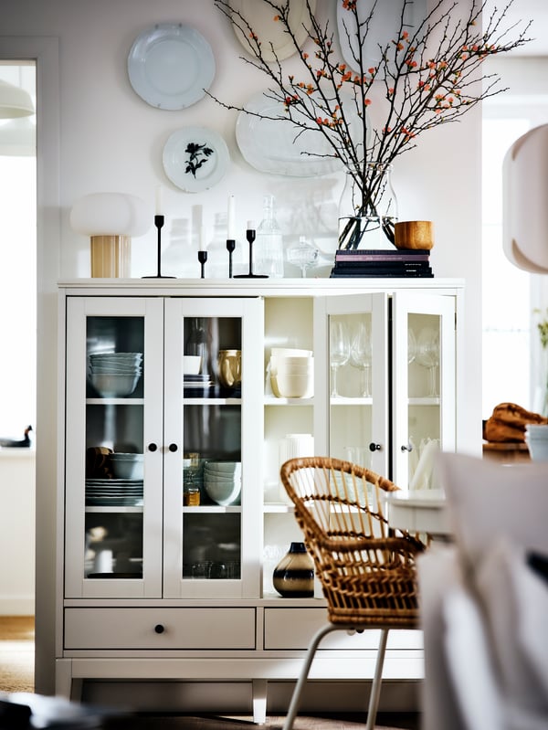 A white IDANÄS cabinet with bi-fold glass doors in a white country-style room filled with objects, by a NILSOVE armchair.