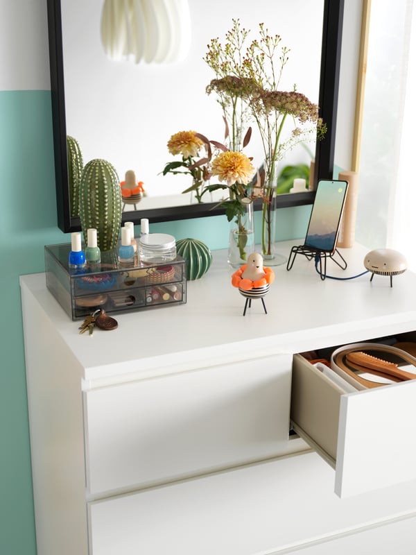 A MALM chest of 6 drawers with one open drawer and many objects on top stands under a NISSEDAL mirror attached to the wall.