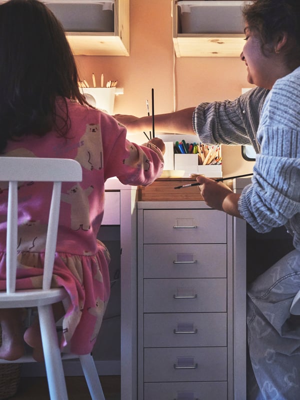 Inspiration för hur man får till två pysselhörnor i ett delat barnrum. En vit HELMER hurts på hjul står mellan två skrivbord där två barn sitter och pysslar.