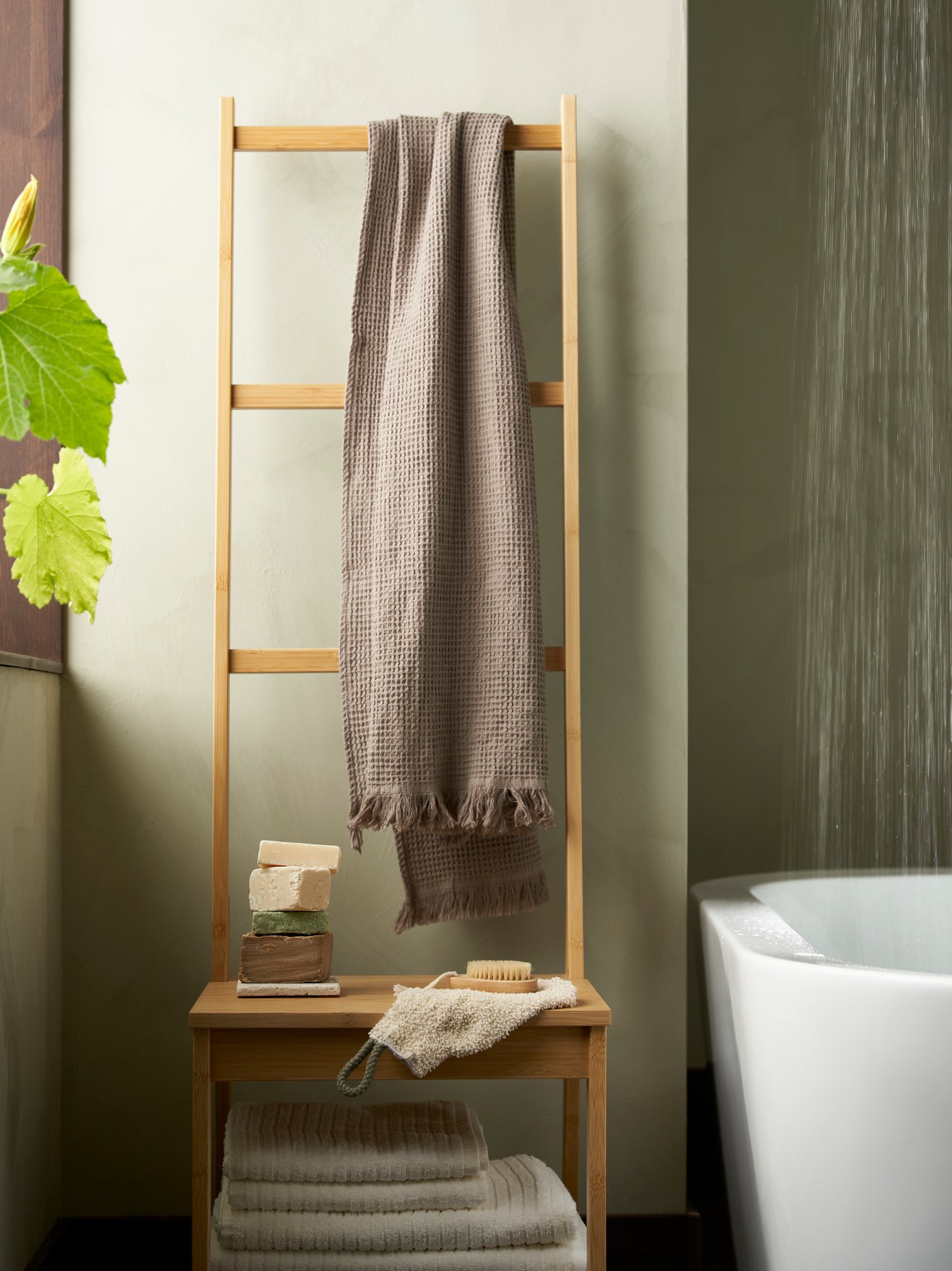 A RÅGRUND towel rack chair in natural bamboo. It is standing next to a bathtub and in front of a light green wall.