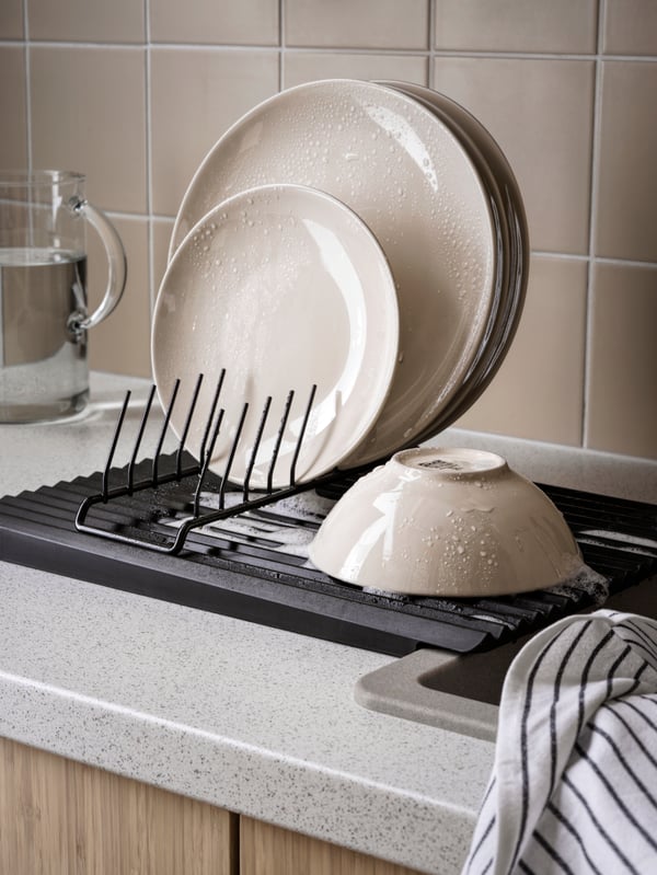 A kitchen worktop with a RINNIG dish drainer and plate holder with plates and a bowl, a RINNIG tea towel and a glass jug.