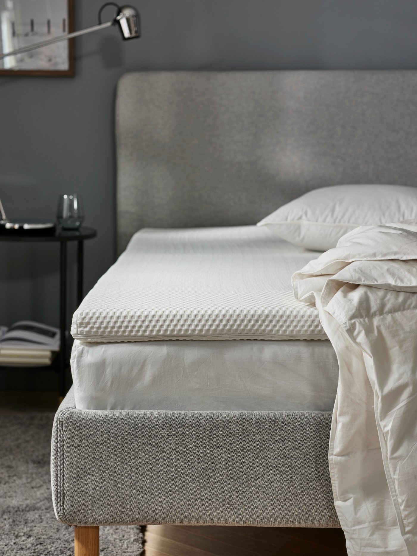 The picture taken from the foot-end of a beige upholstered bed in a bedroom, with the mattress and mattress pad visible.