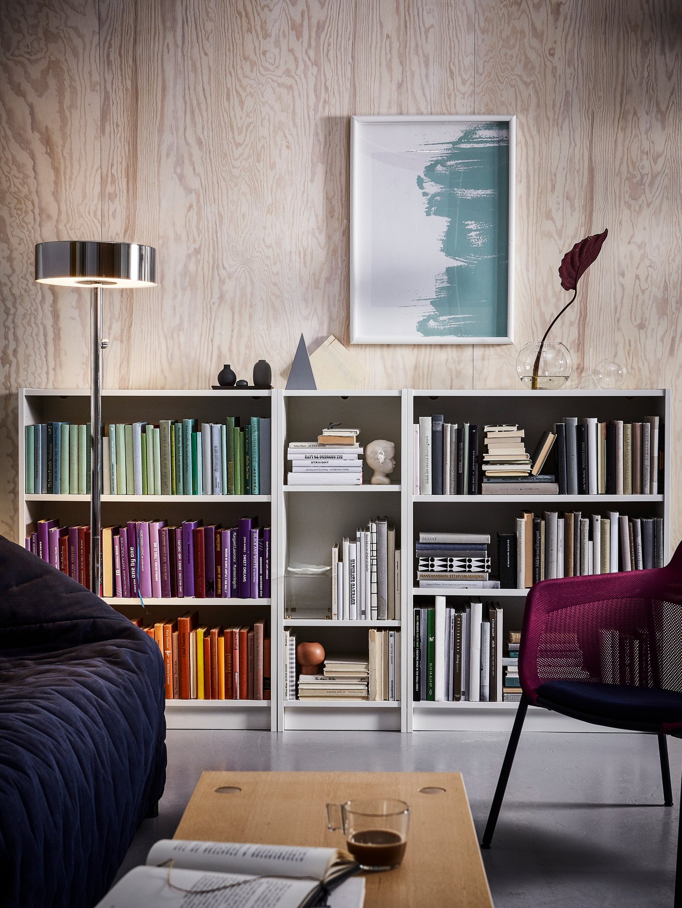 Three BILLY bookcases in a pink living room filled with coloured books. A navy blue sofa and a purple chair are in front.
