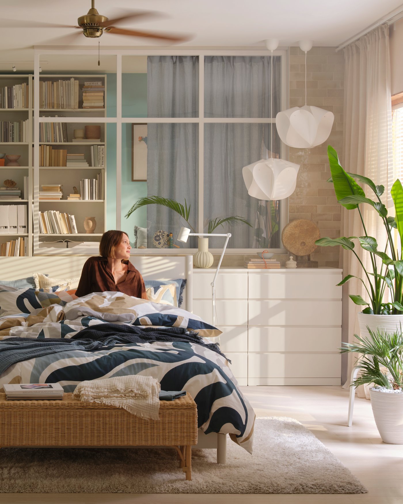 A bright bedroom with white MALM chests of drawers and BILLY bookshelves, a woman sits in a TONSTAD bed gazing out through the window.