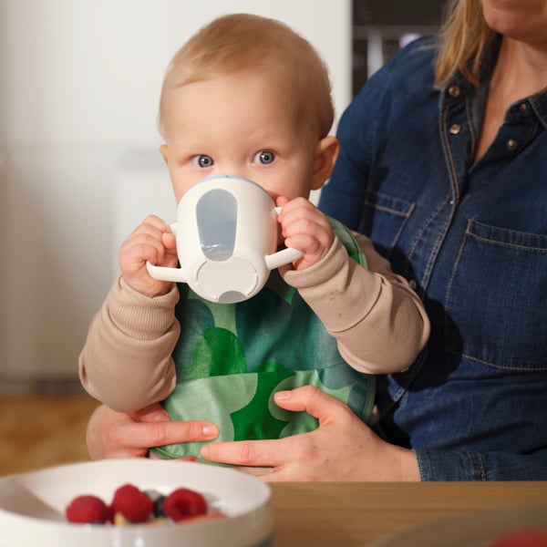 Assis sur les genoux de l’un de ses parents, muni d’un bavoir GRÖNFINK, un enfant boit dans un gobelet BÖRJA. Une assiette est posée sur la table.
