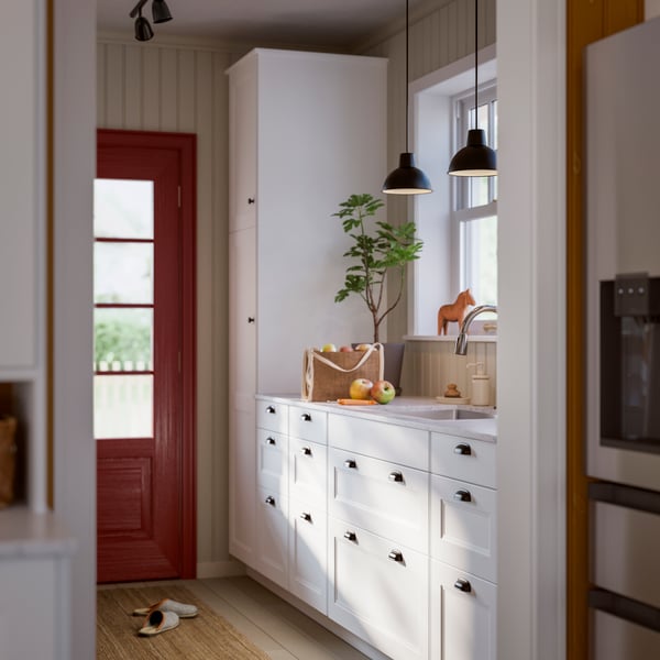 Un vestibule lumineux avec des armoires blanches, une porte en bois et des suspensions noires, qui met en valeur une plante verte et des fruits frais.