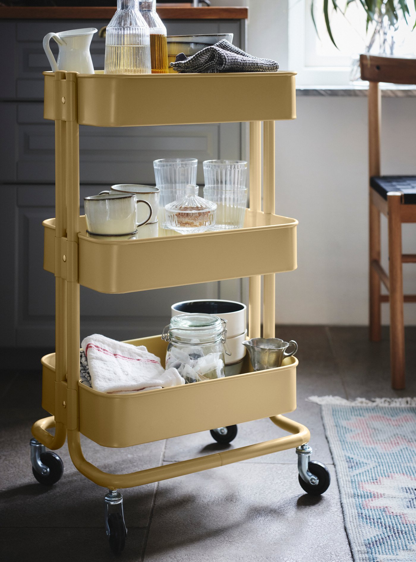 Un chariot de cuisine jaune à trois niveaux est rempli de bouteilles, verres, tasses, bocaux et linges, placé à côté d'une chaise près de la fenêtre.