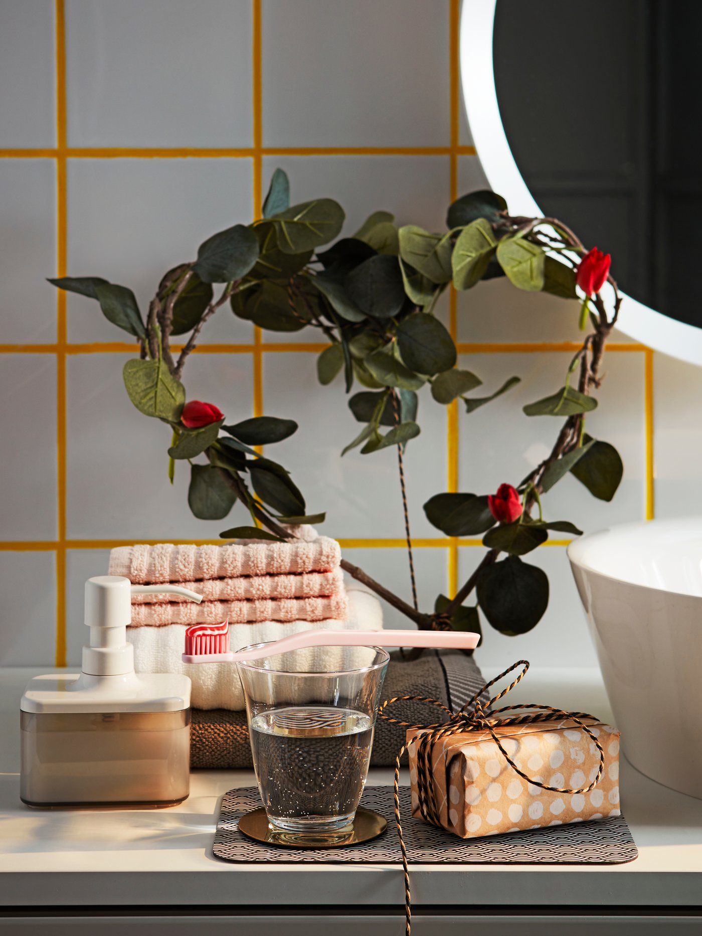 A bathroom shelf with a heart-shaped foliage, folded towels, a toothbrush, a soap dispenser, an IKEA FRAMTRÄDA glass water glass and a small wrapped gift.
