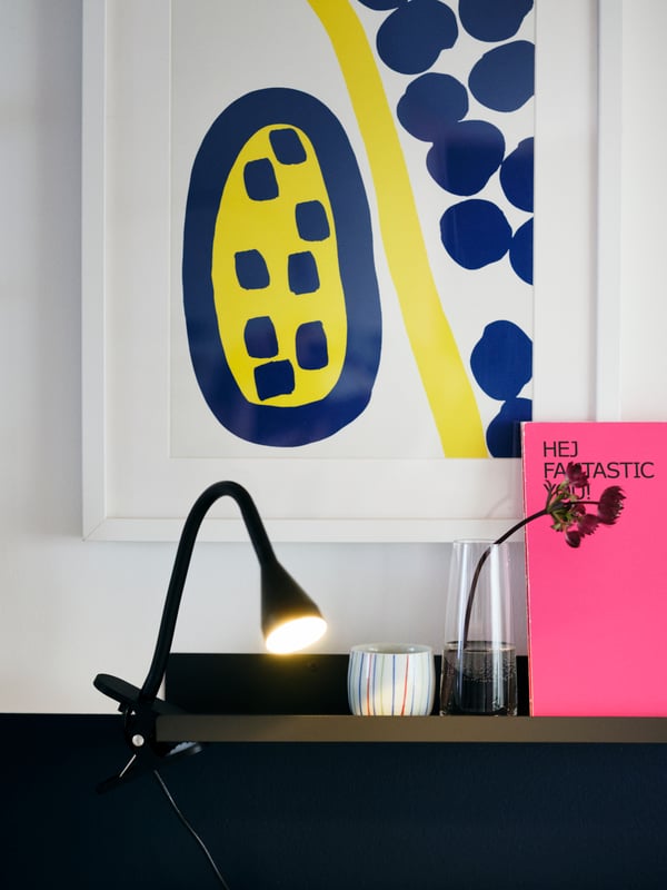 Une applique/spot à pince noir allumé sur une étagère foncée, entourée d'œuvres d’art, de livres et de décorations.