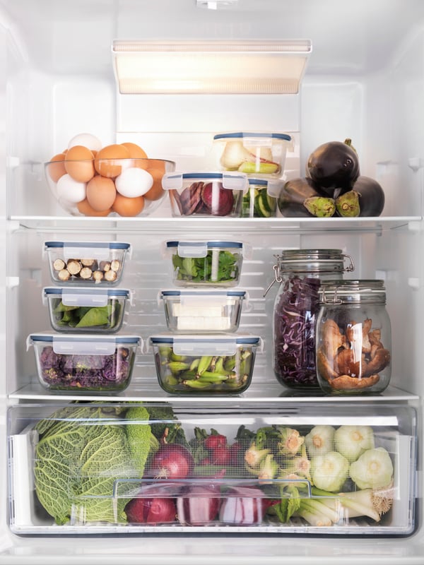 Vista frontal de un refrigerador ordenado con recipientes de vidrio con tapa IKEA 365+ y KORKEN que contienen alimentos frescos. Hay también una fuente BLANDA transparente con huevos y un cajón lleno de verduras en la parte inferior.