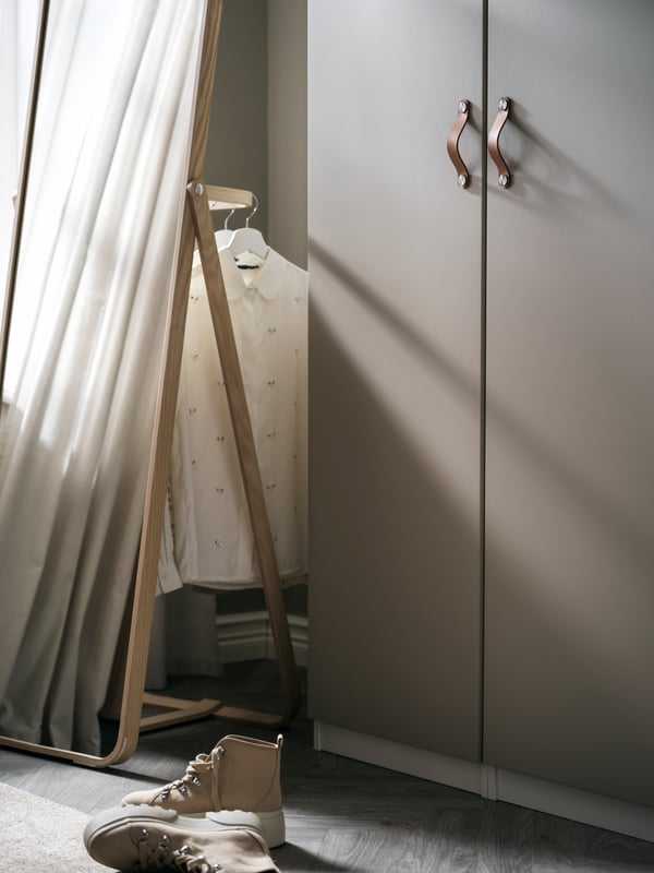 A corner with an ash IKORNNES mirror holding clothes on hangers next to a PAX wardrobe with grey-beige REINSVOLL doors.