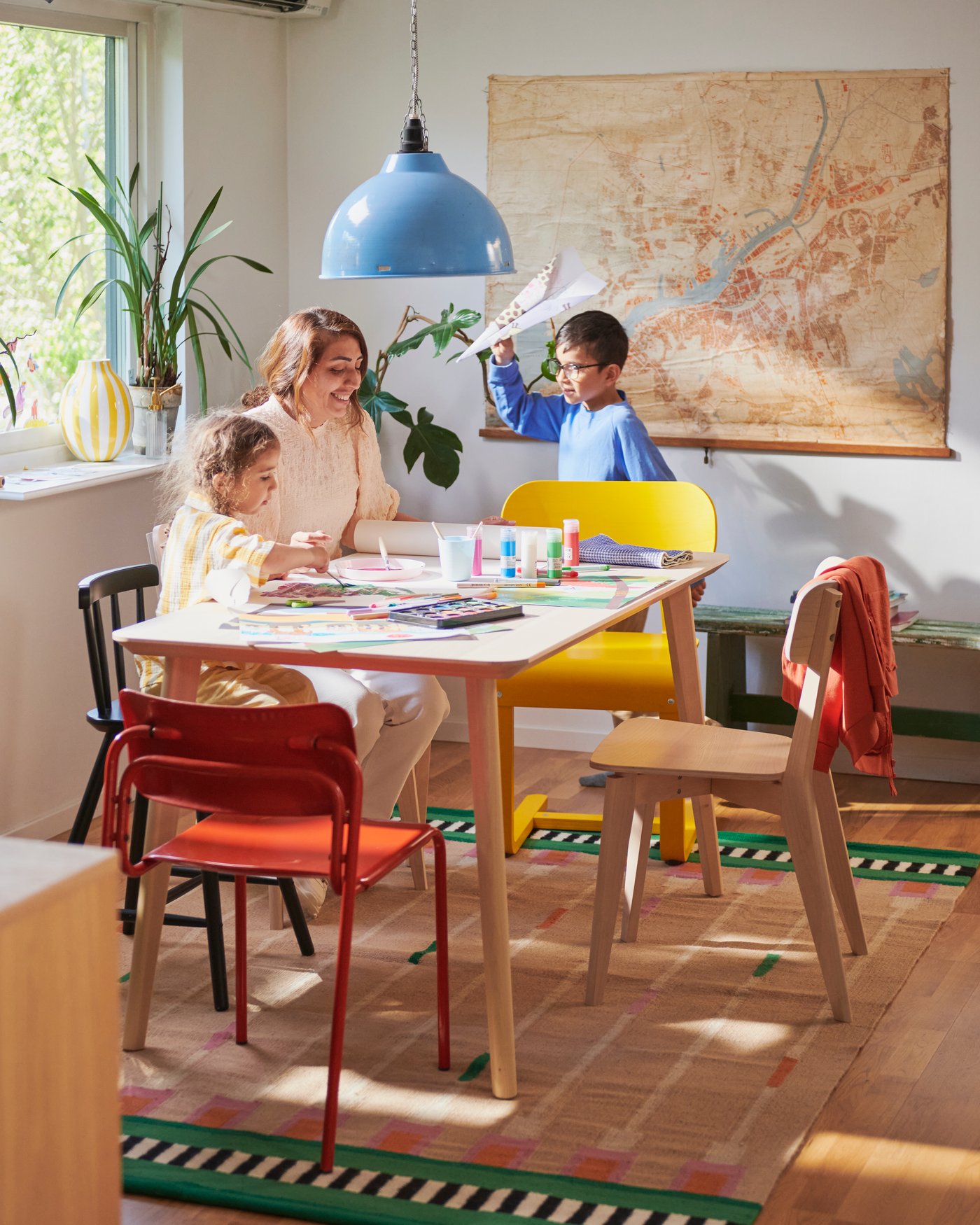 Heller Essbereich mit einem Tisch und bunten Stühlen, an dem mehrere Personen sitzen und malen; im Hintergrund eine große Landkarte, eine blaue Hängeleuchte und Tageslicht, das durch das Fenster fällt.