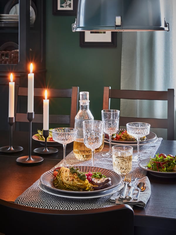 A black table with lit candles, a black/white table-runner, dinner for two and a vintage-looking glassware series.