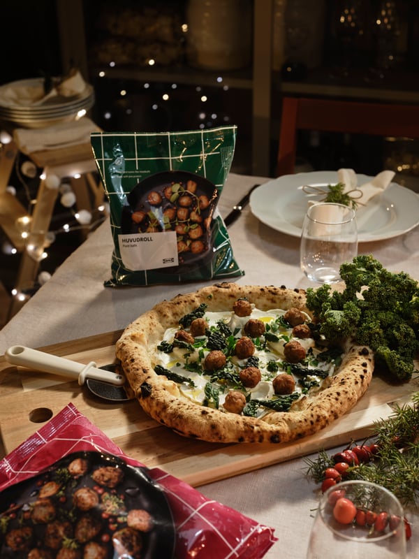 Bord med pizza toppad med köttbullar och grönt, på träbräda. I bakgrunden syns HUVUDROLL påse, tallrikar, glas och ljusslingor.