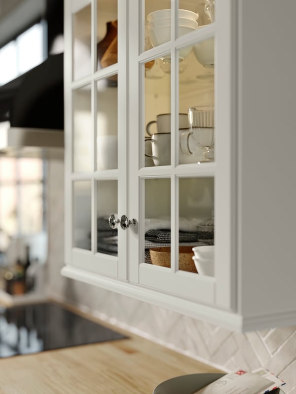 A cabinet with BODBYN glass doors in off-white with shelves holding GLADELIG mugs in gray and VARDAGEN bowls in off-white.