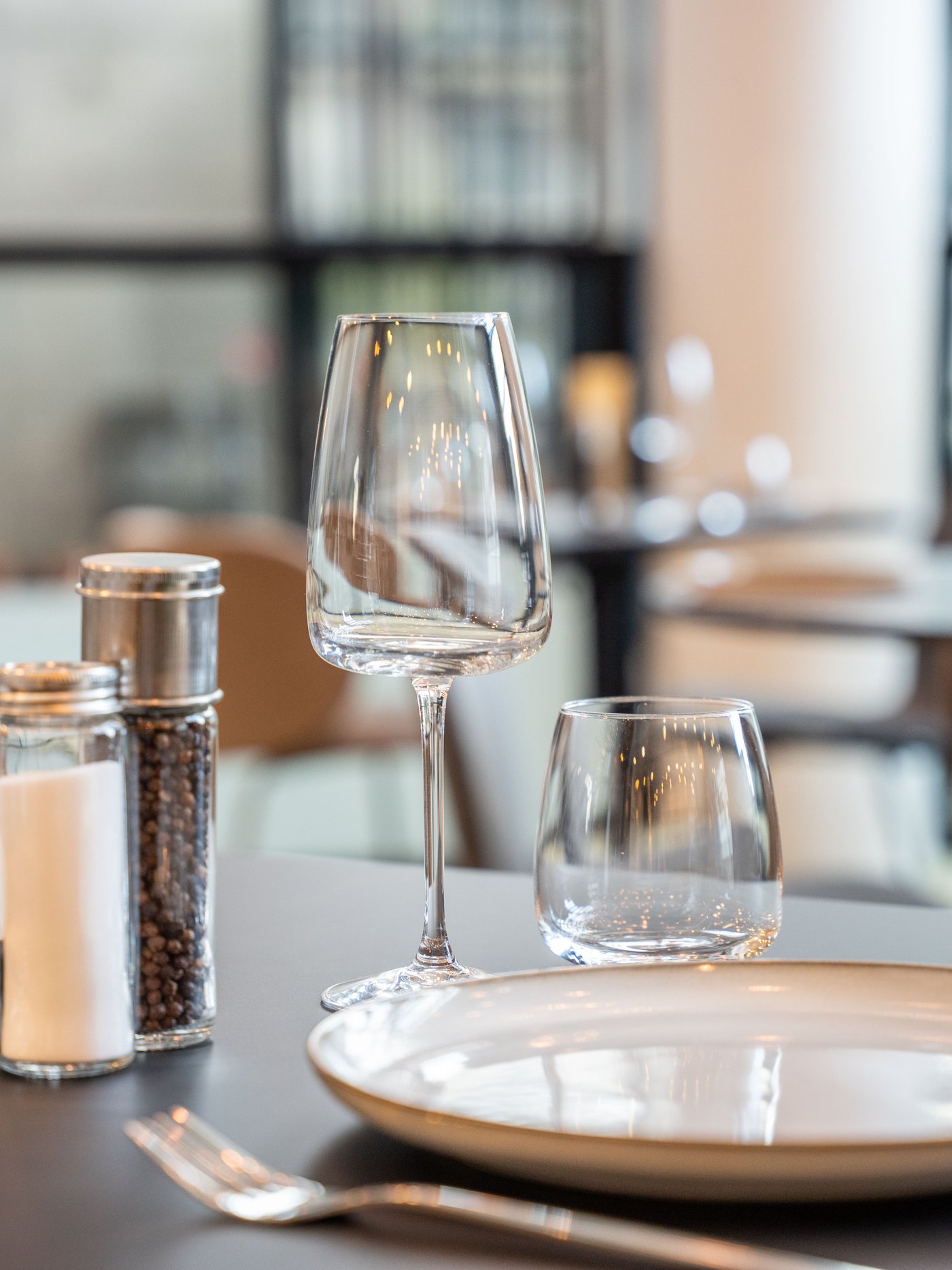 Wit bord, glazen en peper-zoutstel op gedekte tafel in stijlvol restaurant. Zachte belichting en moderne sfeer met zicht op andere tafels.