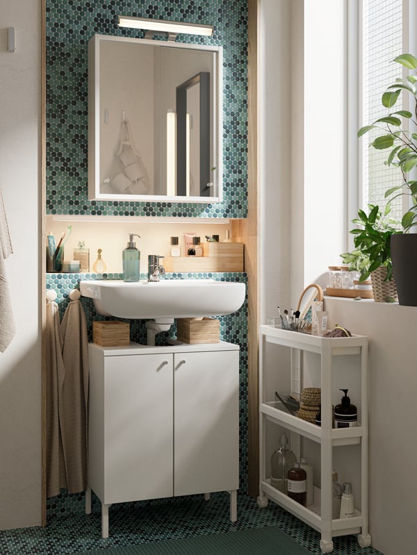 A green-tiled bathroom, white bathroom furniture set, white trolley and plants on a windowsill.