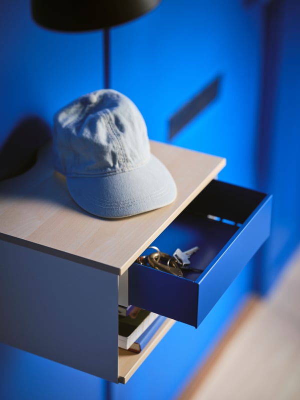 A birch effect white/blue STOMSÖ wall-mounted beside table on a blue wall with a hat on top and the drawer open showing keys.