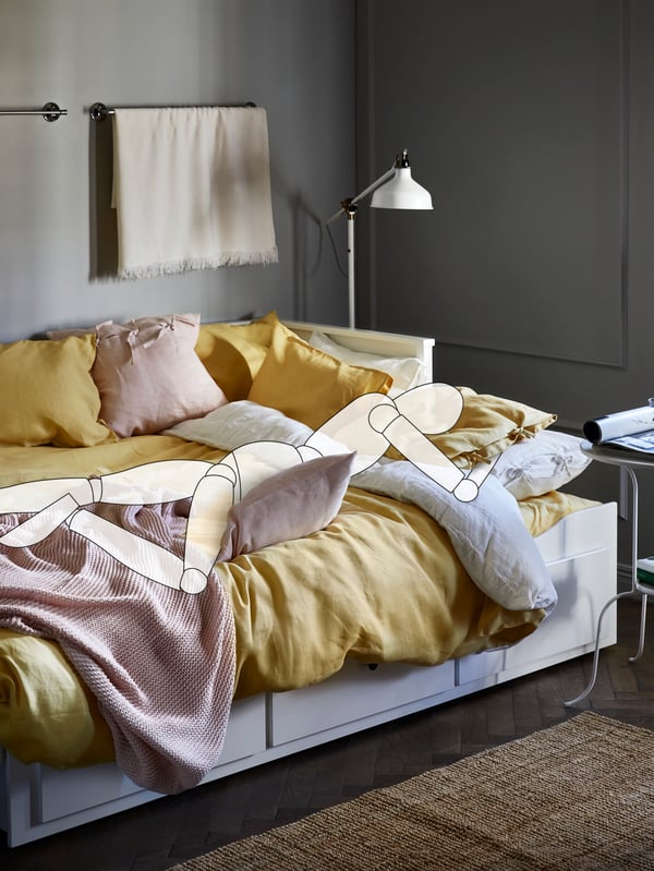 A cartoon drawing of a person sleeps on a HEMNES day-bed with yellow bed linen and a pink throw, with a lamp above the bed.