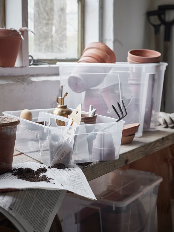 A rustic, wooden worktop scattered with pots and planting accessories, many of them in transparent SAMLA storage boxes.