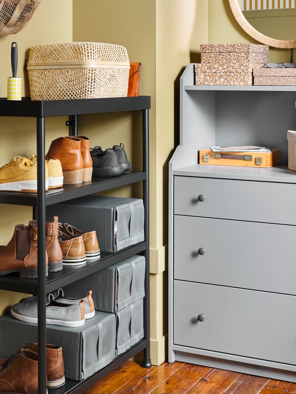 A black KORNSJÖ shelving unit with shoes and shoe boxes and a grey HAUGA chest of drawers stand in a corner in a hall.